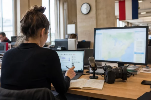 Journaliste dans une rédaction parisienne préparant un sujet sur un déplacement diplomatique, avec caméra et écrans flous.