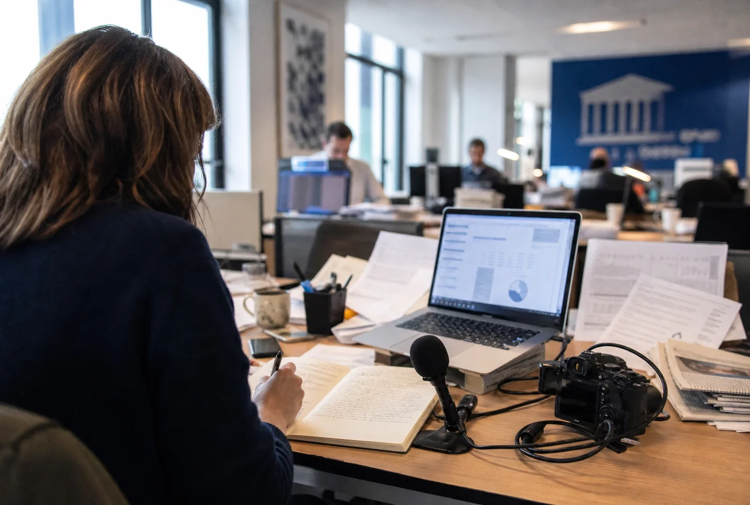 Dans une rédaction française, un journaliste prépare une analyse politique avec micro, carnet et écran flou.