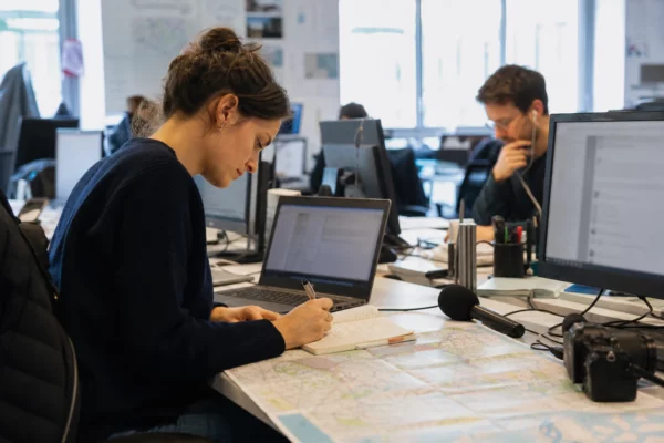Journaliste dans une rédaction française préparant un sujet politique avec carnet, micro et écrans flous.