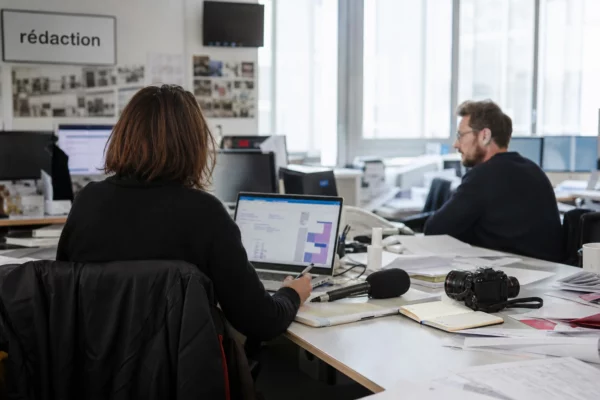 Journalistes dans une rédaction française devant écrans flous, micro sans logo et carnet ouvert