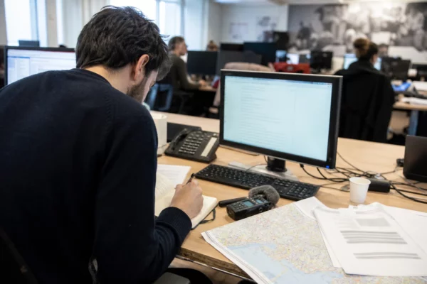 Journaliste en rédaction préparant un sujet sur la loi Ripost, avec carnet, micro et écrans flous.