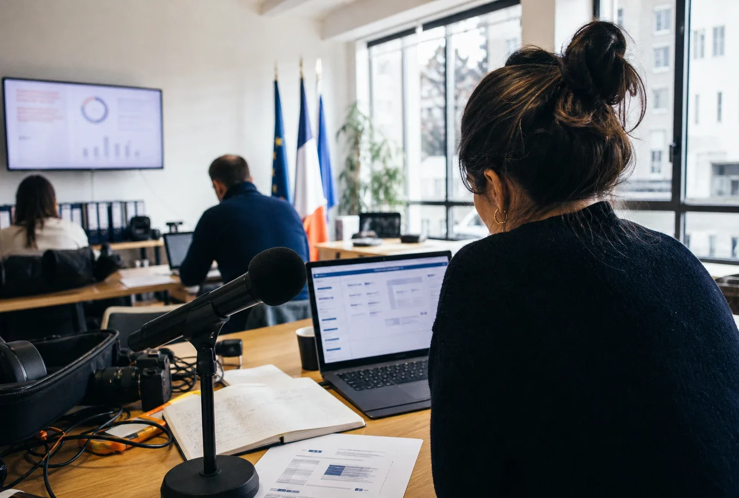 Journaliste en rédaction préparant un sujet politique avec micro, écran flou et carnet ouvert.