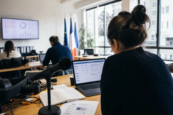 Journaliste en rédaction préparant un sujet politique avec micro, écran flou et carnet ouvert.