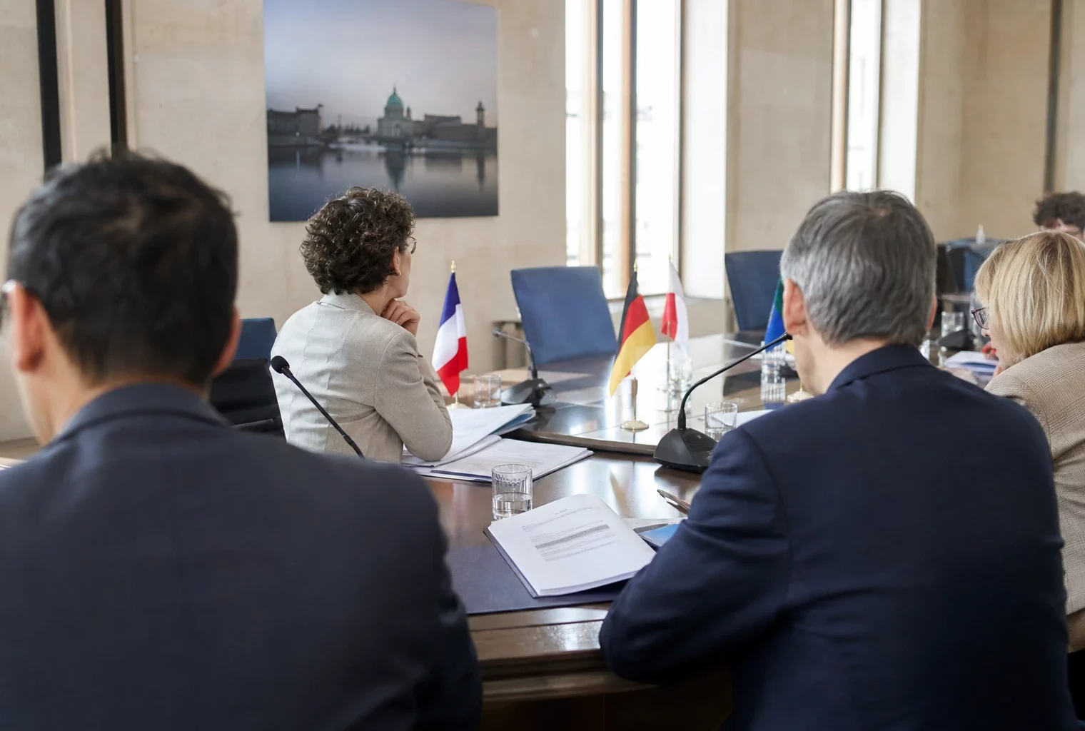 Réunion politique européenne sobre avec silhouettes de dos, dossiers ouverts et micros sur une table