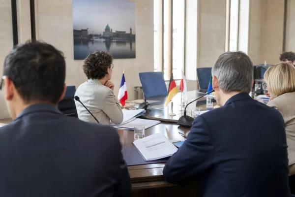 Réunion politique européenne sobre avec silhouettes de dos, dossiers ouverts et micros sur une table