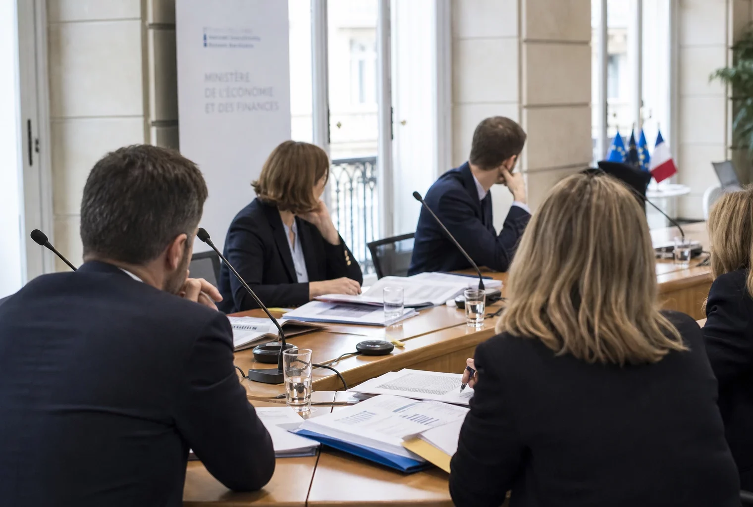 Réunion européenne sobre avec dossiers, micros et silhouettes de dos dans une salle claire