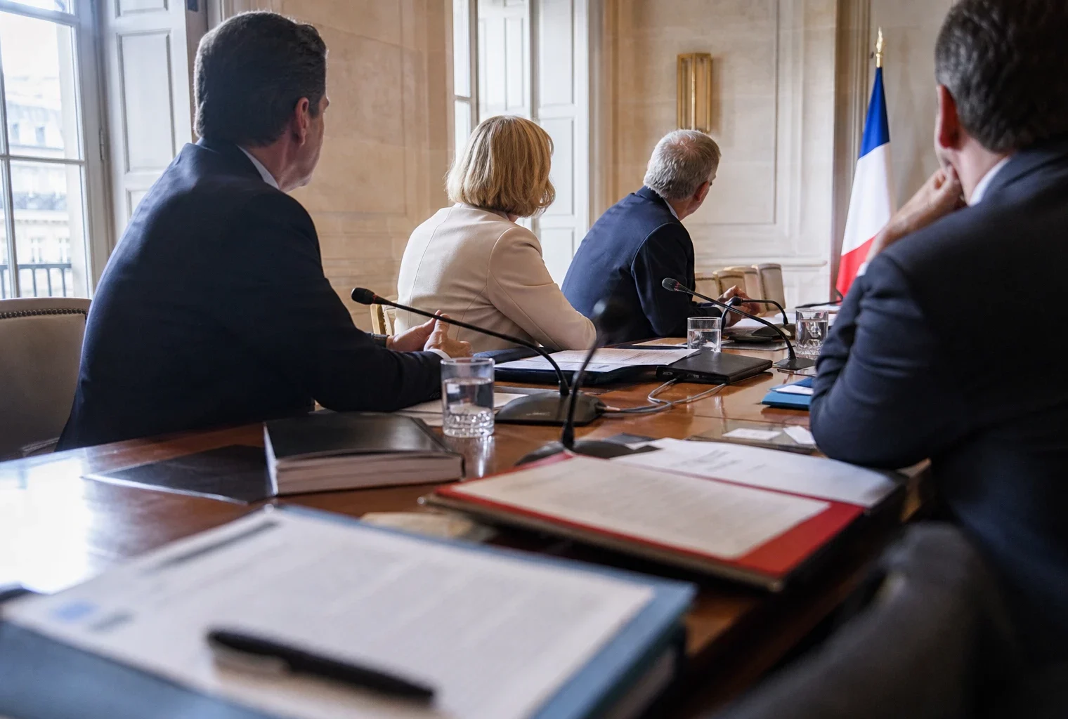 Réunion européenne sobre avec silhouettes de dos, dossiers ouverts et drapeaux de table discrets dans une salle lumineuse.