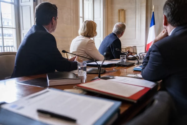 Réunion européenne sobre avec silhouettes de dos, dossiers ouverts et drapeaux de table discrets dans une salle lumineuse.