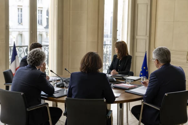 Réunion européenne sobre avec cinq silhouettes de dos, dossiers et petits drapeaux de table dans une salle claire.