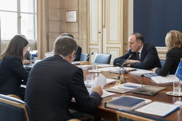 Réunion européenne sobre avec silhouettes anonymes, dossiers et micros sur table, en lumière naturelle claire.