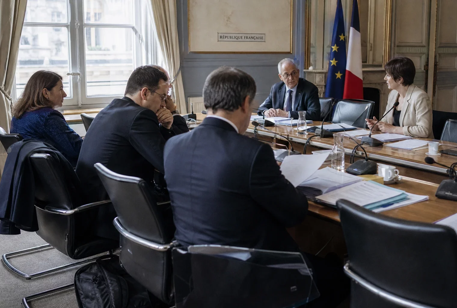 Réunion européenne sobre avec dossiers, silhouettes de dos et drapeaux de table discrets dans une lumière claire