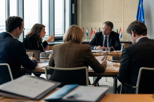 Réunion européenne sobre avec dossiers, micros et silhouettes de dos autour d’une table institutionnelle lumineuse.