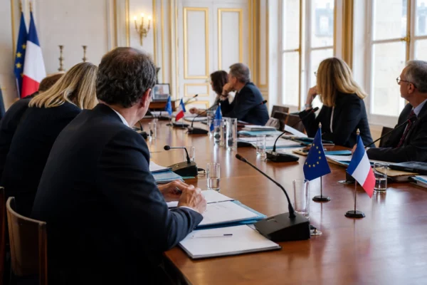 Réunion européenne sobre avec plusieurs silhouettes autour d’une table, dossiers ouverts et lumière naturelle claire.