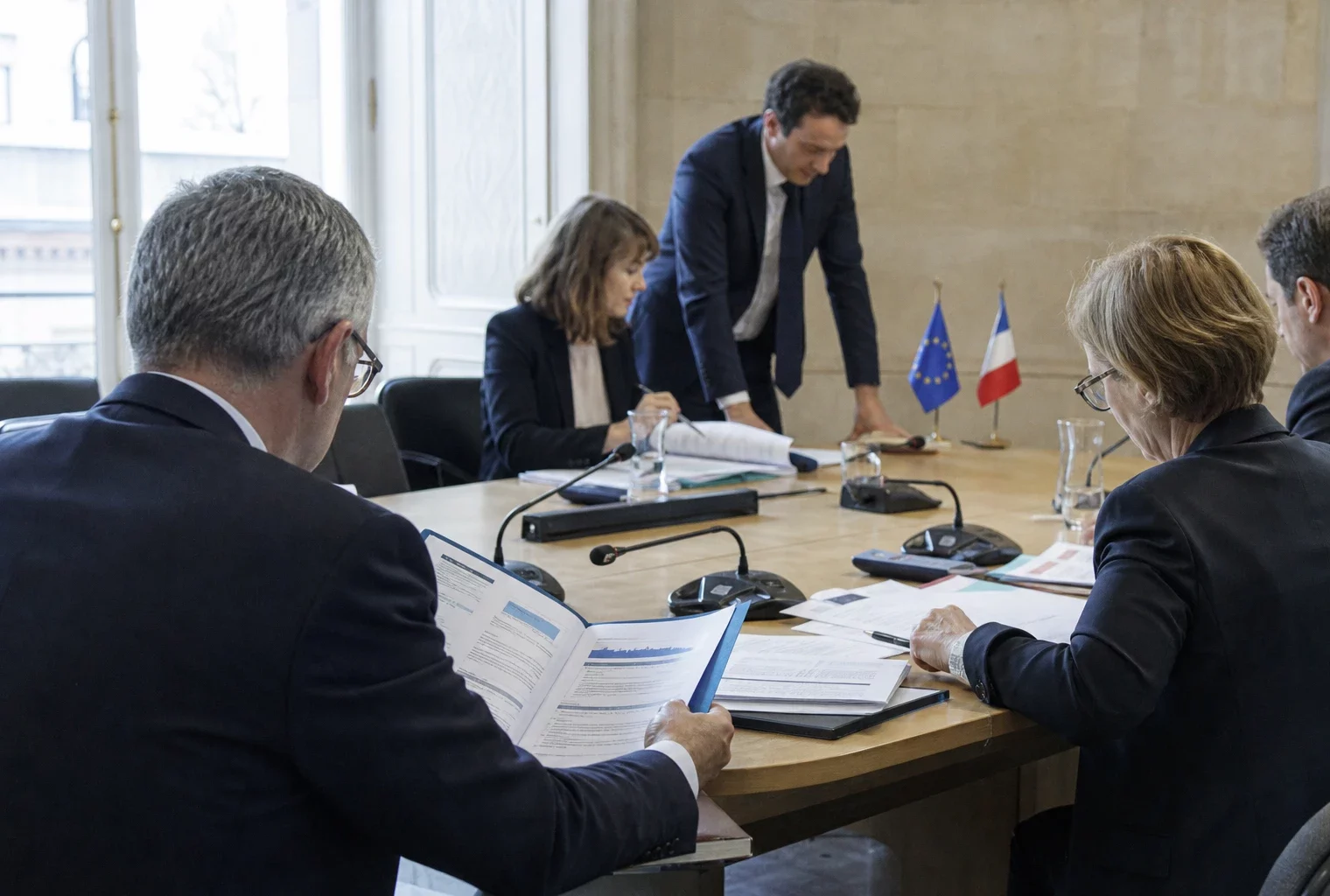 Réunion européenne sobre avec dossiers, micros et silhouettes de dos dans une salle institutionnelle lumineuse.