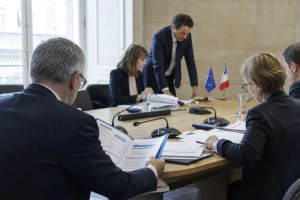 Réunion européenne sobre avec dossiers, micros et silhouettes de dos dans une salle institutionnelle lumineuse.