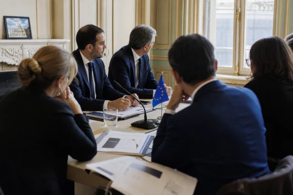 Réunion européenne sobre avec plusieurs silhouettes de dos, dossiers ouverts et petits drapeaux de table dans une lumière claire.