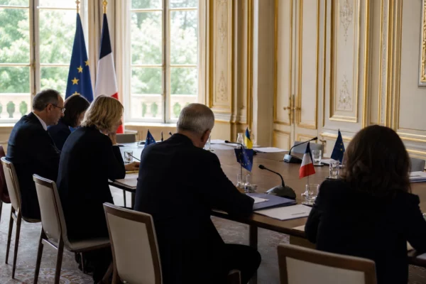 Réunion européenne sobre avec silhouettes de dos, dossiers ouverts et petits drapeaux de table dans une lumière claire.