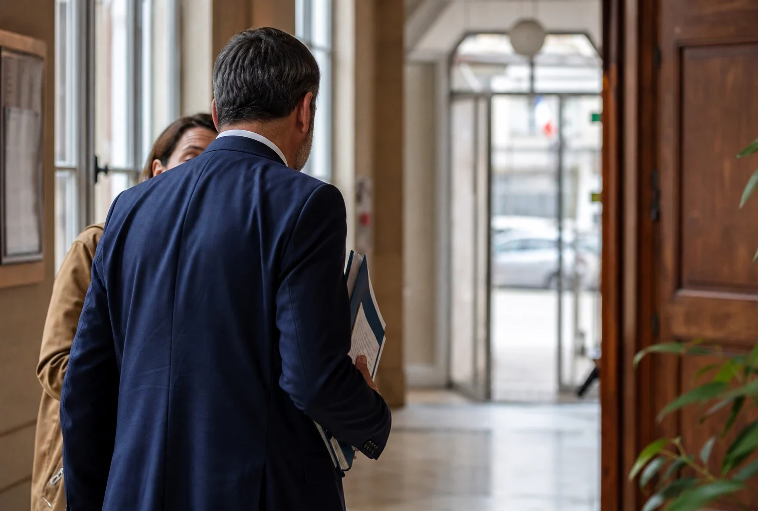 Un élu local anonyme dans un hall de mairie, dossier en main, lumière naturelle et arrière-plan urbain flou.