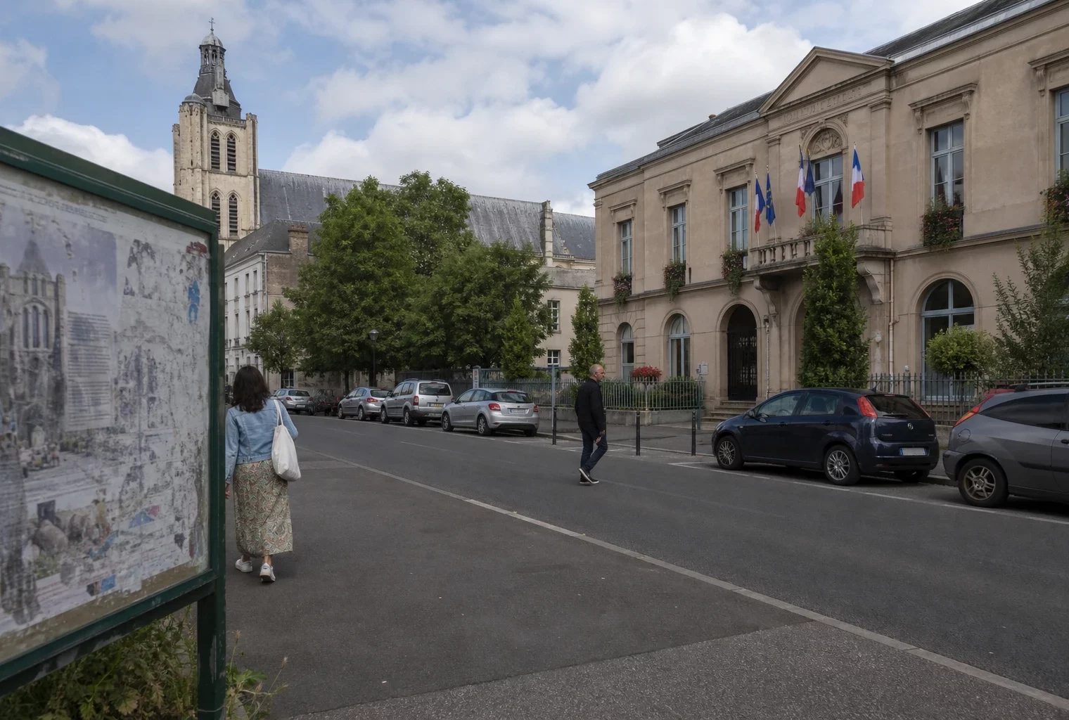 Rue de Saint-Denis près de la mairie, avec passants anonymes et un rappel discret du chantier de la basilique.