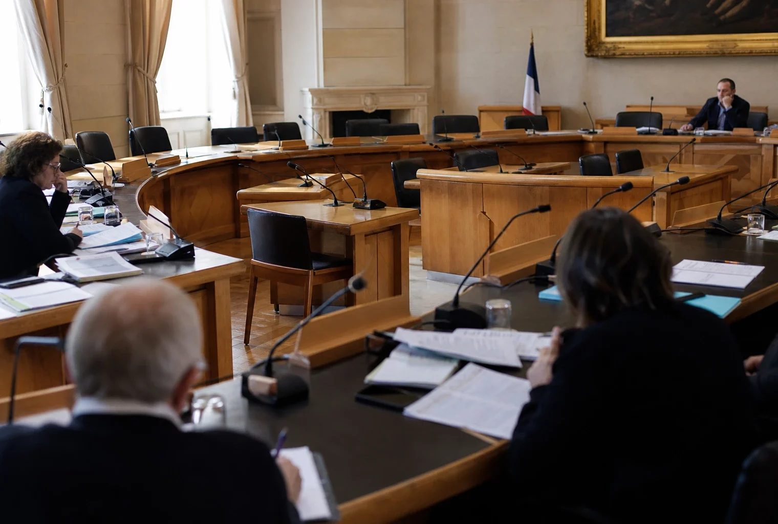 Salle de conseil municipal vide avec micros et dossiers flous, sous une lumière naturelle claire