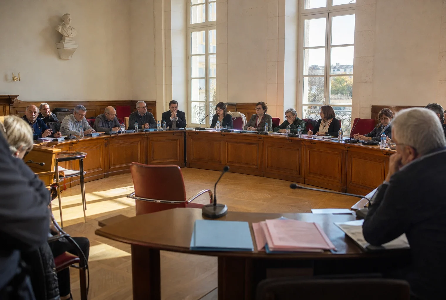 Salle municipale lumineuse avec chaise vide, micros et dossiers flous, ambiance politique tendue en arrière-plan.