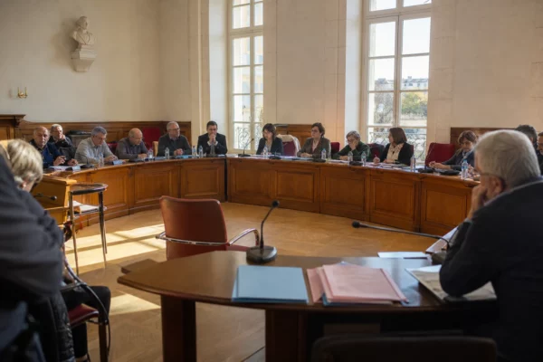 Salle municipale lumineuse avec chaise vide, micros et dossiers flous, ambiance politique tendue en arrière-plan.