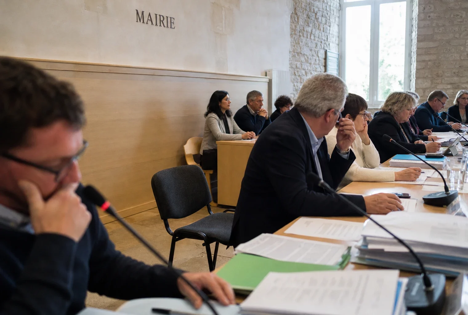 Salle municipale française avec chaise vide, micros et dossiers flous, ambiance de conseil local en lumière naturelle.