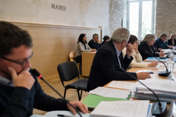 Salle municipale française avec chaise vide, micros et dossiers flous, ambiance de conseil local en lumière naturelle.