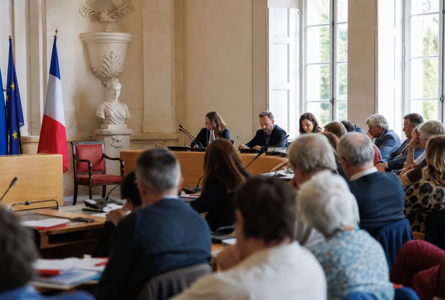 Salle municipale claire avec chaise vide subtile, micros et dossiers flous autour d’une réunion locale.