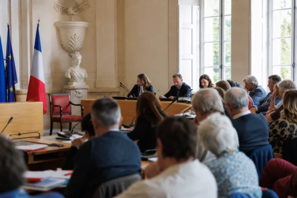 Salle municipale claire avec chaise vide subtile, micros et dossiers flous autour d’une réunion locale.