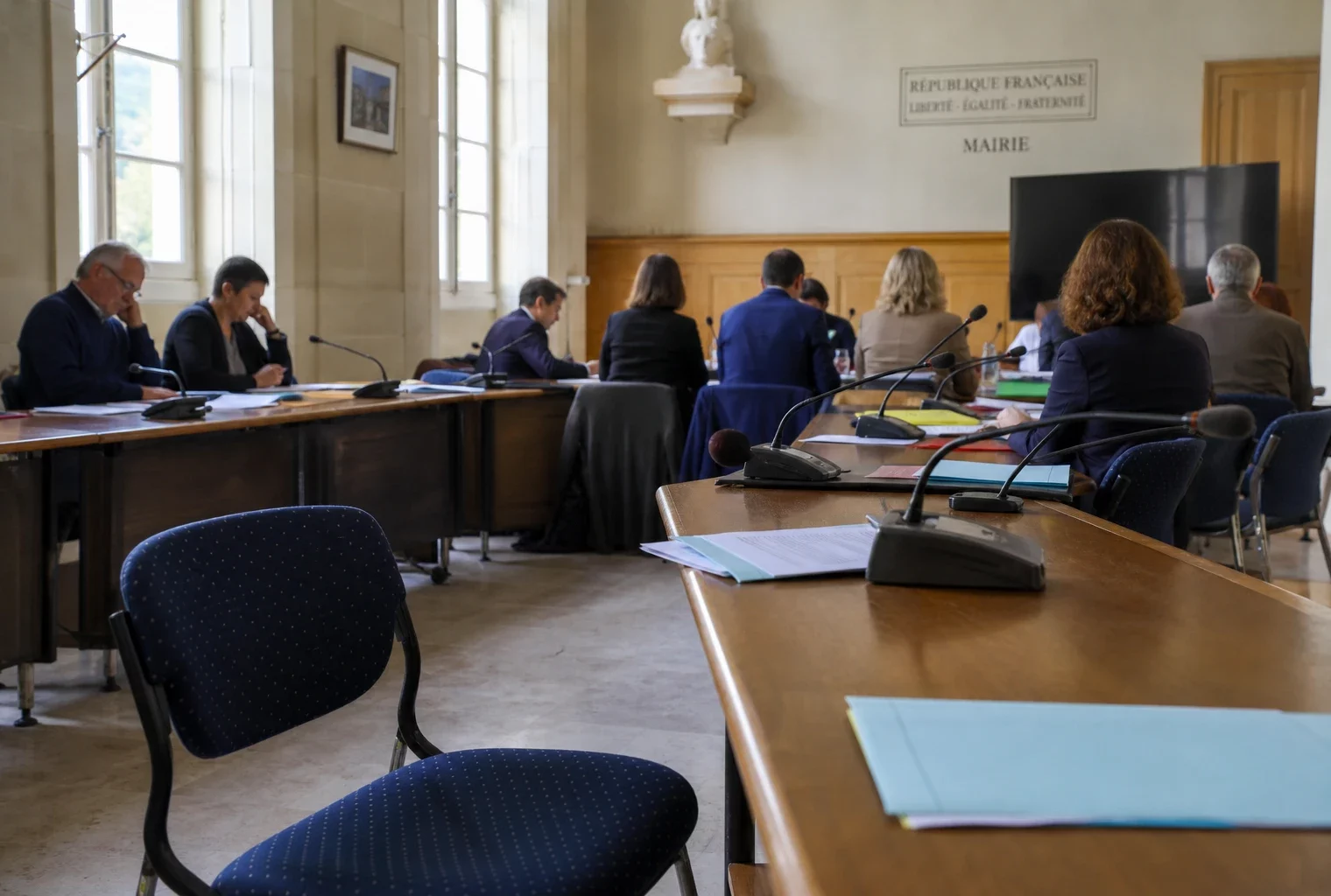 Salle municipale lumineuse avec chaise vide, micros et dossiers flous dans une ambiance politique calme.