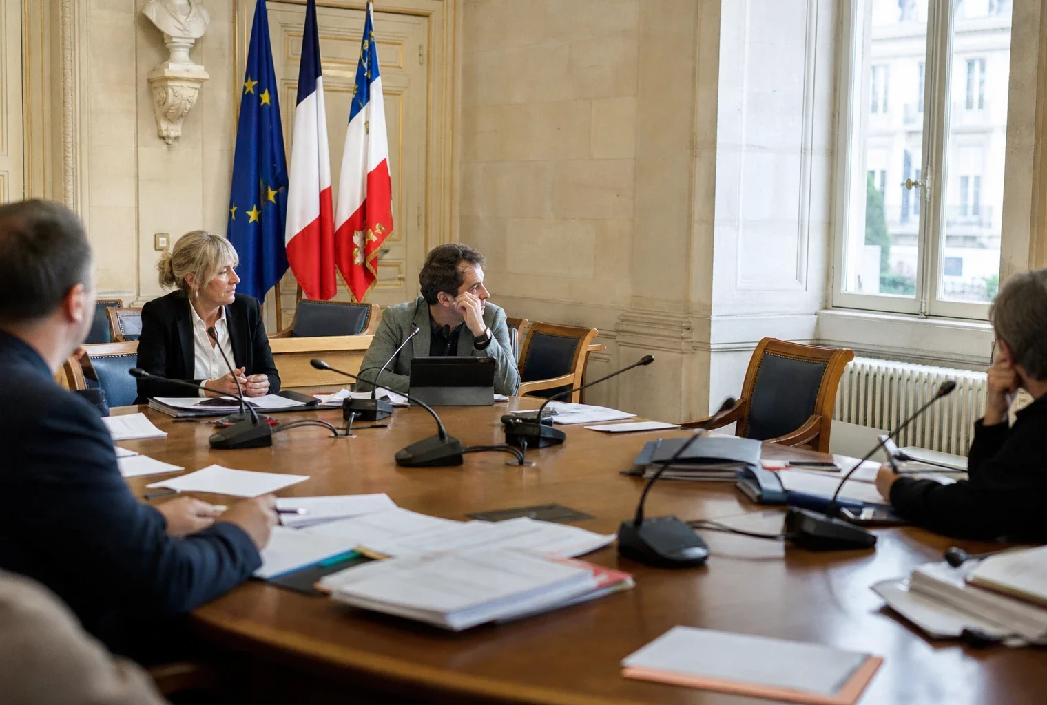 Salle municipale lumineuse avec micros, dossiers flous et chaise vide autour d’une table de conseil.