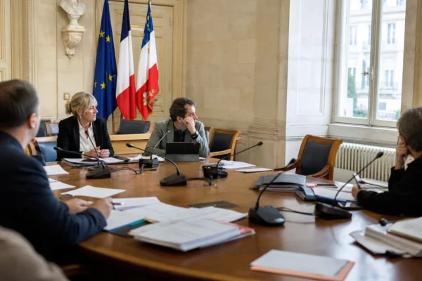 Salle municipale lumineuse avec micros, dossiers flous et chaise vide autour d’une table de conseil.