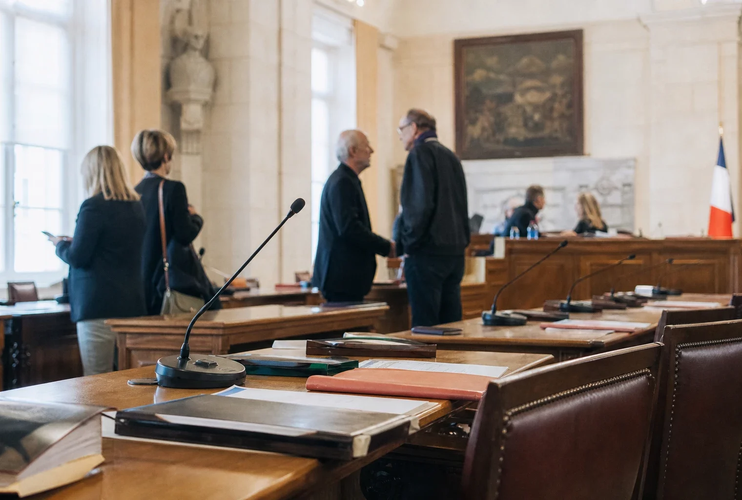 Salle municipale éclairée naturellement avec micros, dossiers flous et chaise vide lors d’un conseil local.