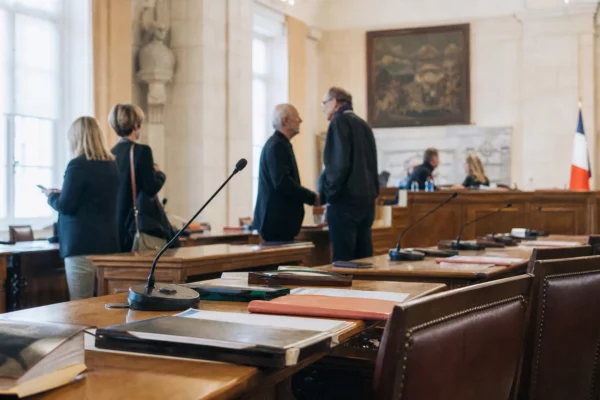 Salle municipale éclairée naturellement avec micros, dossiers flous et chaise vide lors d’un conseil local.