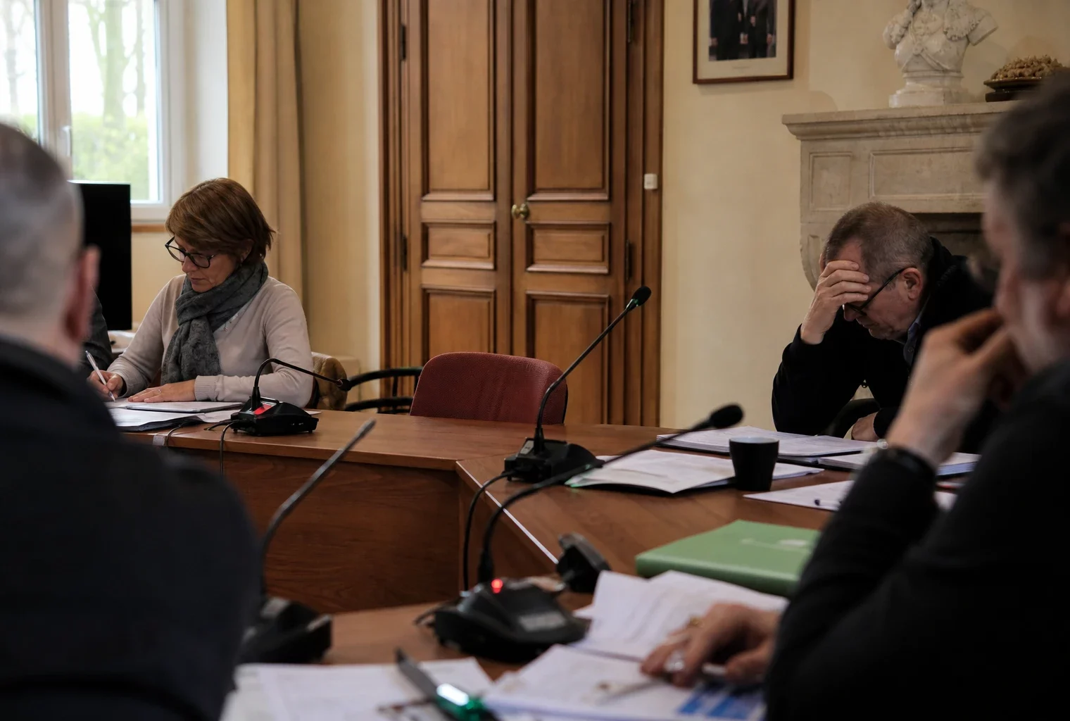 Salle municipale girondine avec micros, dossiers flous et chaise vide lors d’une réunion de crise.