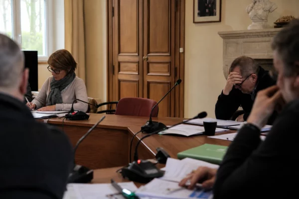 Salle municipale girondine avec micros, dossiers flous et chaise vide lors d’une réunion de crise.