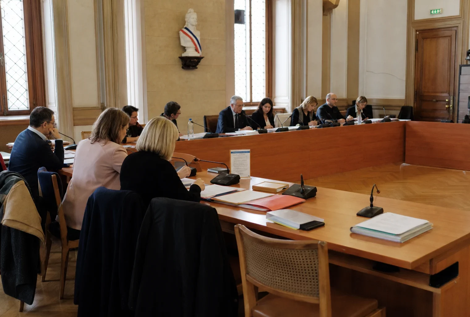 Salle municipale lumineuse avec micros, dossiers flous et chaise vide lors d’une réunion locale sur le carburant