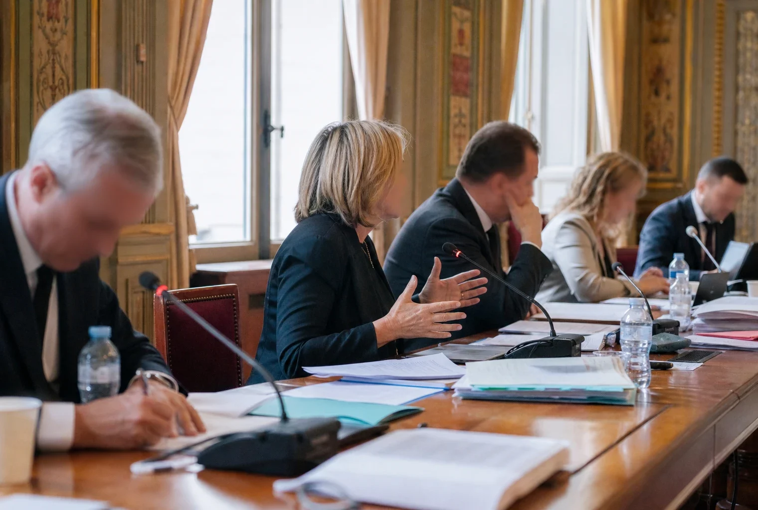 Réunion de commission parlementaire au Sénat avec micros, dossiers et silhouettes anonymes en lumière naturelle.