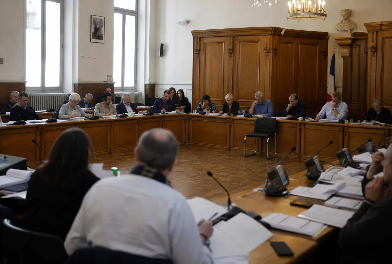 Salle municipale en réunion avec chaise vide, micros et dossiers flous dans une scène claire de conseil local.
