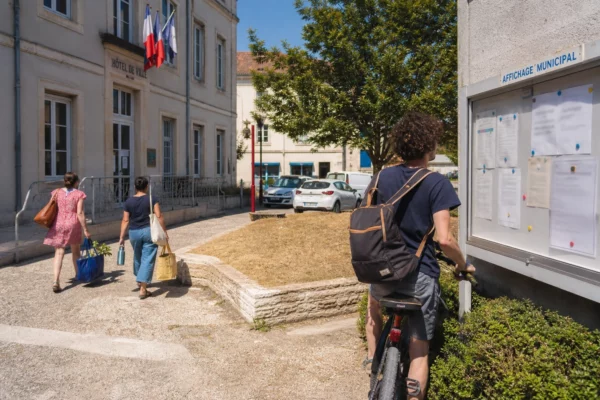 Forecourt municipal dans une ville moyenne française, avec habitants anonymes et atmosphère de restrictions d’eau.