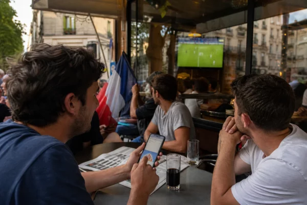 Supporters de football dans un café parisien, l’un consulte un smartphone tandis qu’une télévision diffuse le tournoi.