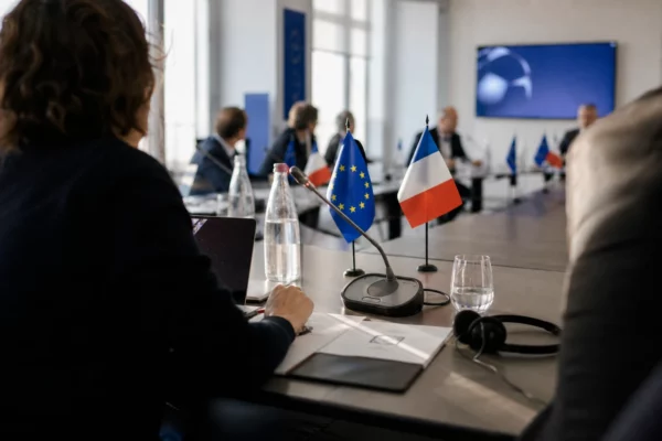 Table diplomatique européenne avec dossiers, écouteurs, verres d’eau et petits drapeaux discrets dans une salle institutionnelle.