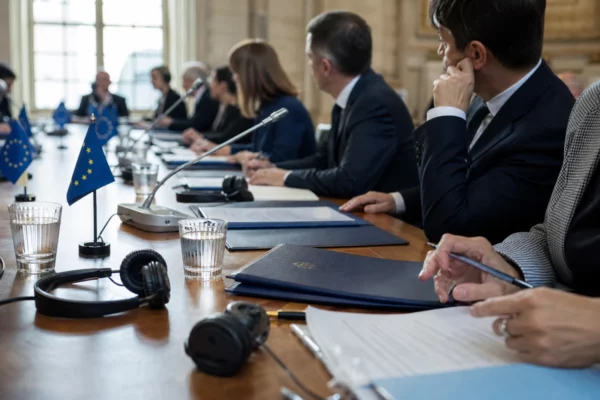 Table de négociation européenne avec dossiers, écouteurs et verres d’eau, autour de silhouettes anonymes.