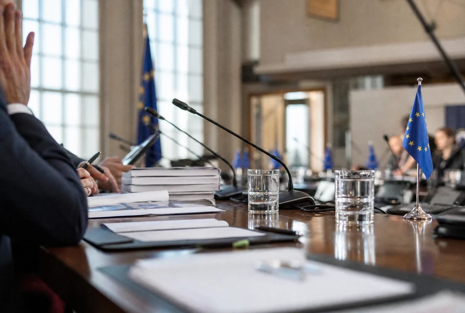 Table diplomatique européenne avec dossiers, micros, écouteurs, verres d’eau et petits drapeaux UE dans une salle claire