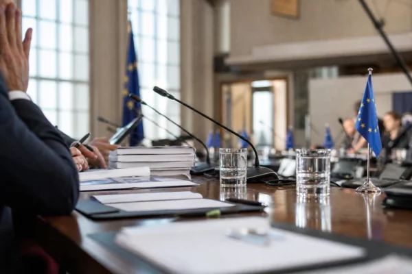 Table diplomatique européenne avec dossiers, micros, écouteurs, verres d’eau et petits drapeaux UE dans une salle claire