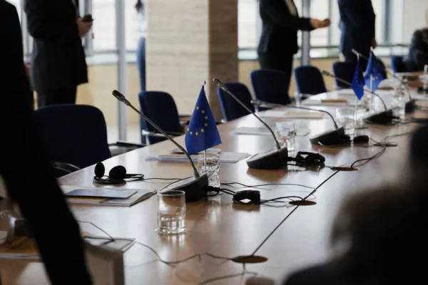 Table diplomatique européenne avec dossiers, casques et drapeaux miniatures, en réunion institutionnelle lumineuse.