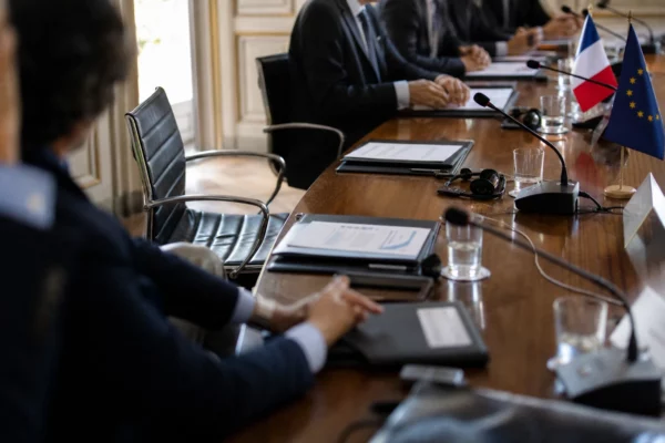 Table de négociation avec micros, dossiers fermés et silhouettes anonymes dans une salle lumineuse