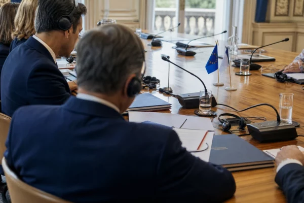 Table de négociation européenne avec micros, dossiers fermés, écouteurs et silhouettes anonymes en salle claire.