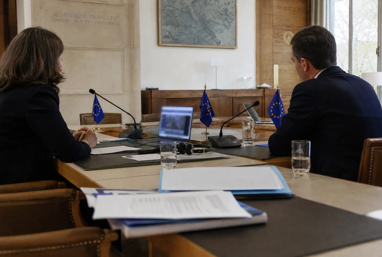 Table de négociation institutionnelle avec micros, dossiers fermés, écouteurs et silhouettes anonymes dans une salle claire.